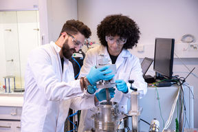 Two people in laboratory working with research equipment wearing safety goggles.