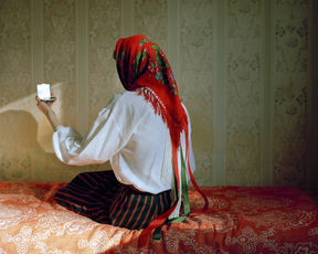 woman in white shirt and red printed headscarf sites on a bed with her back turned, looking in a small square handmirror