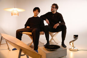 Two men dressed in dark clothes sit in the middle of furniture they have designed