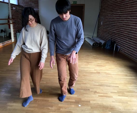 Image from a performative workshop. Two participants walk side-by-side in a indoor space