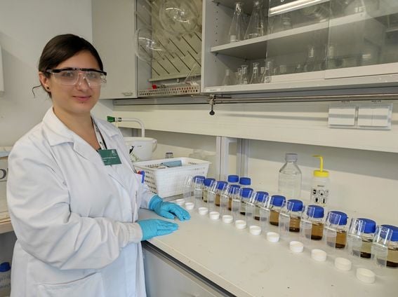 Color photo of a scientist wearing a white lab coat and protective glasses stands at a lab bench with sample containers lined up in clear glass, plastic and blue lids
