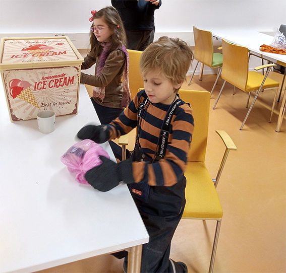 Two children with pink plastic bags containing ice cream mixture sitting at tables