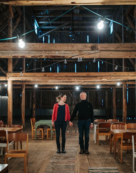 Arnevi Rautanen ja Juha Utriainen seisovat hymyillen käsikädessä maalaistalon ladon ylisillä, joka on sisustettu räsymatoin ja koristevaloin. Tilassa on myös useita pöytäryhmiä tuoleineen.