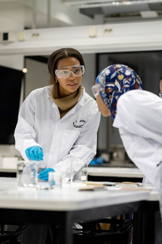 Kaksi suojalaseihin, valkoisiin laboratoriotakkeihin ja sinisiin suojakäsineisiin pukeutunutta yläkoululaista tarkastelee mittalasissa olevaa näytettä.