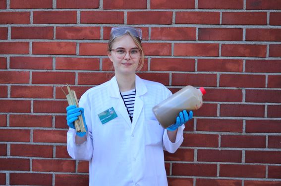Color photo of young researcher standing in front of a red brick wall, wearing a lab coat and protective glasses, blue protective gloves, holding plant stems and a large bottle filled with brown liquid