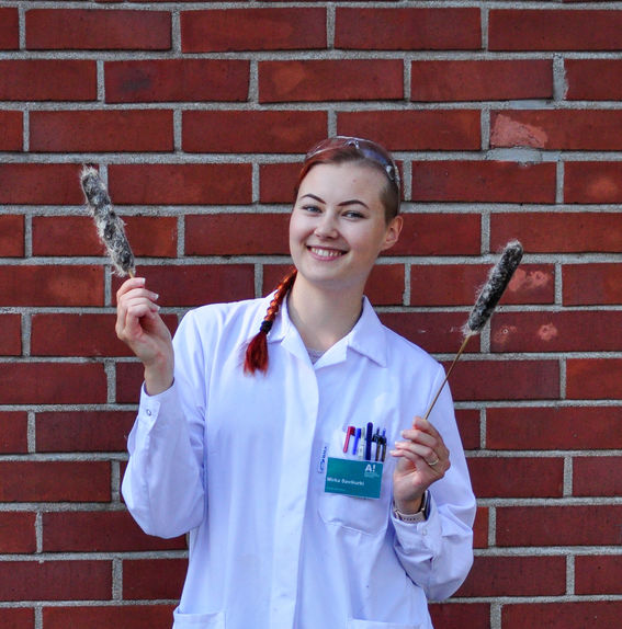 Color photo of a smiling person on a red brick background wearing a white lab coat, protective glasses on their head, and holding a cattail flower in each hand