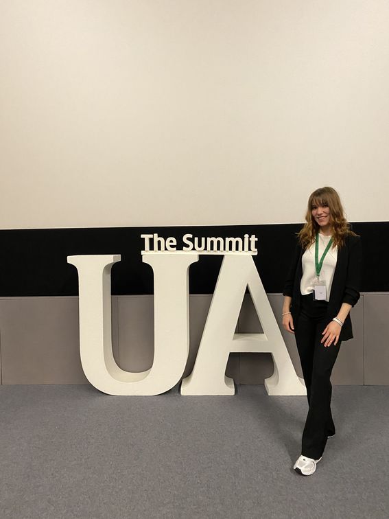 Photo of young woman standing next to a logo reading "UA"