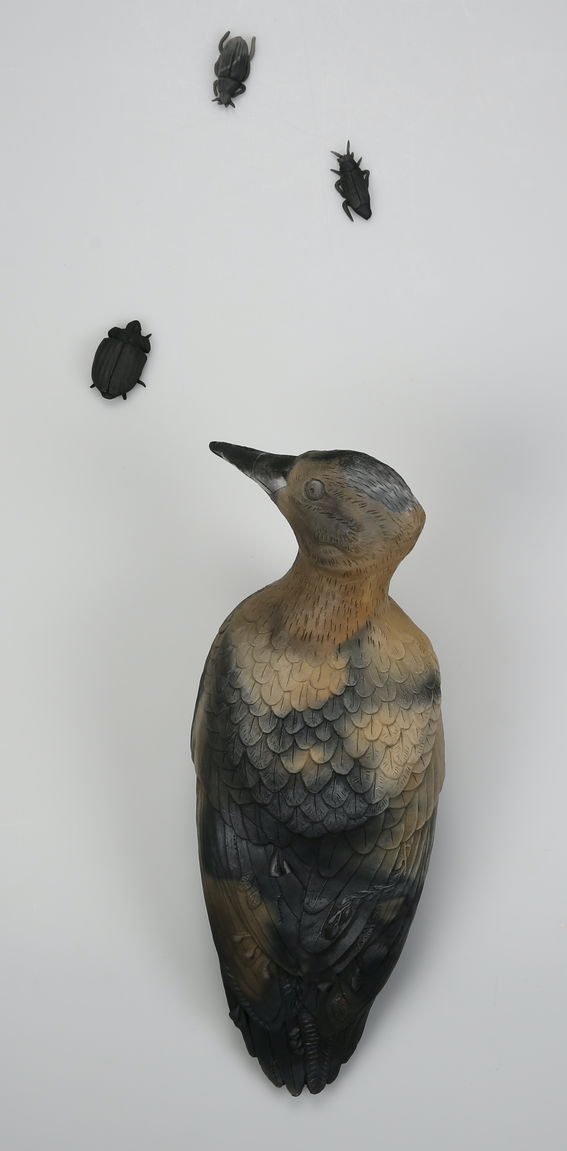A clay sculpture of a woodpecker who is curiously looking at three clay bugs in front of it