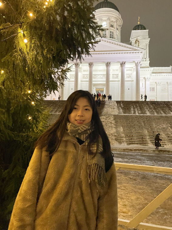A woman is standing next to a big tree with lights on it. A big white church is shown in the background.