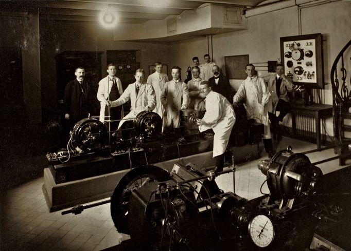 Historiallisessa kuvassa ryhmä miesopiskelijoita ja heidän opettajansa poseeraavat suurten mekaanisten koneiden äärellä