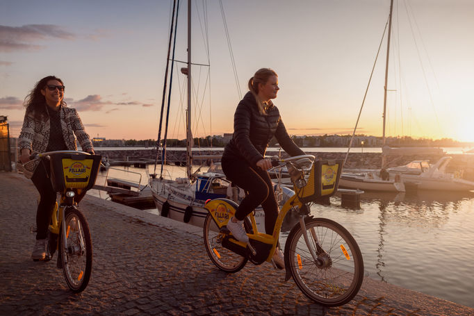 two people riding bicycles close to the seashore with boats in the background