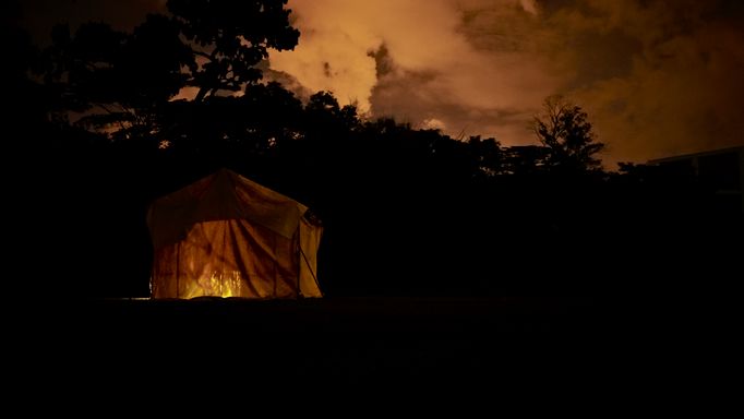 Still from the film Like Shadows Through Leaves, 2021. A lamp-lit tent stands in a dark landscape, it's orange interior matching the orange skyline