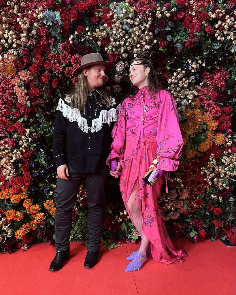 two people smiling at each other holding hands at a red carpet event with a floral wall in the background