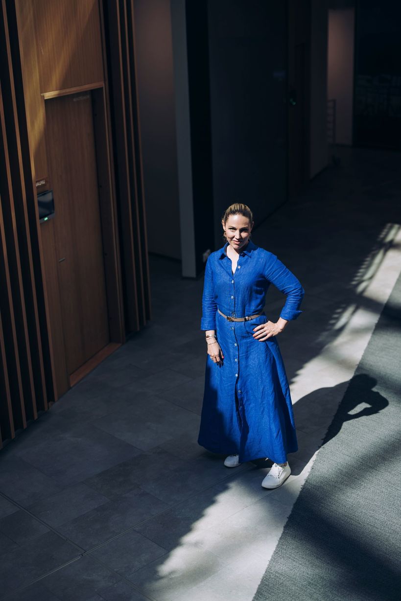 Siniseen farkkumekkoon pukeutunut Maija Jokela seisoo avarassa sisätilassa käytävällä suuren puisen oven vieressä.