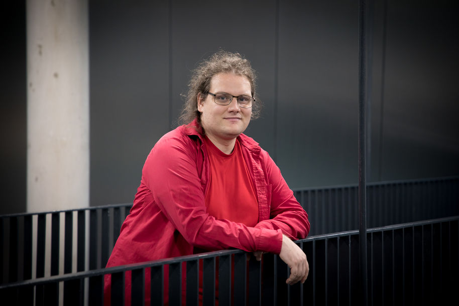 Matti Nelimarkka seisoo ja nojaa kaiteeseen Tietotekniikan talolla. Hänellä on punaiset vaatteet ja taustalla on harmaa seinä ja valkoinen pylväs.