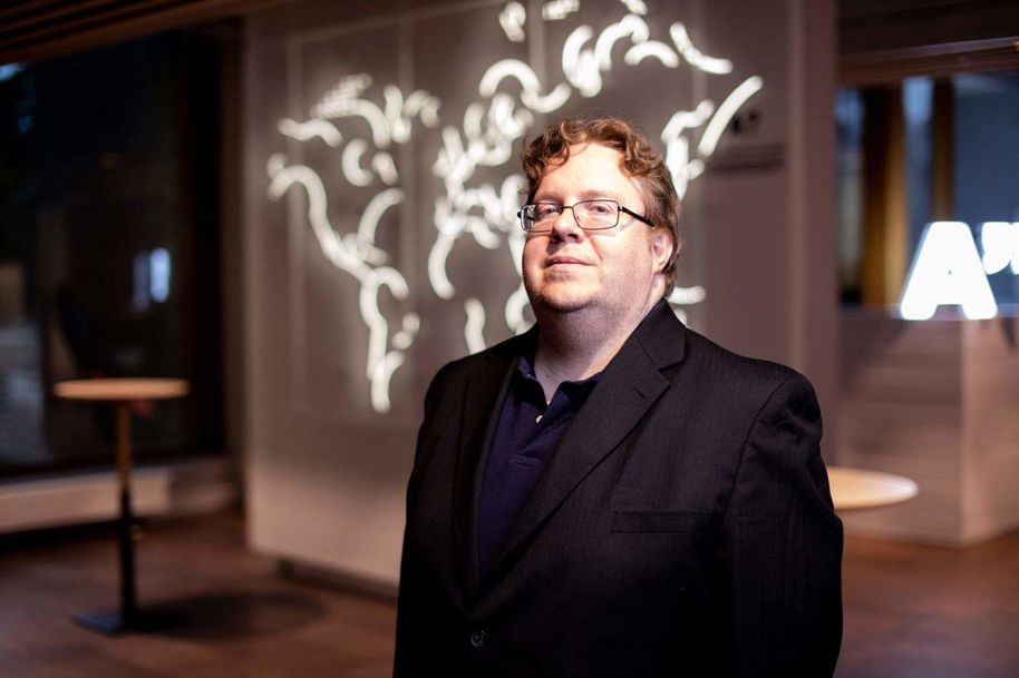 Janne Lindqvist seisoo mustassa puvussa taustallaan Aallon A-kirjainvalotaulu ja valotaideteos seinällä