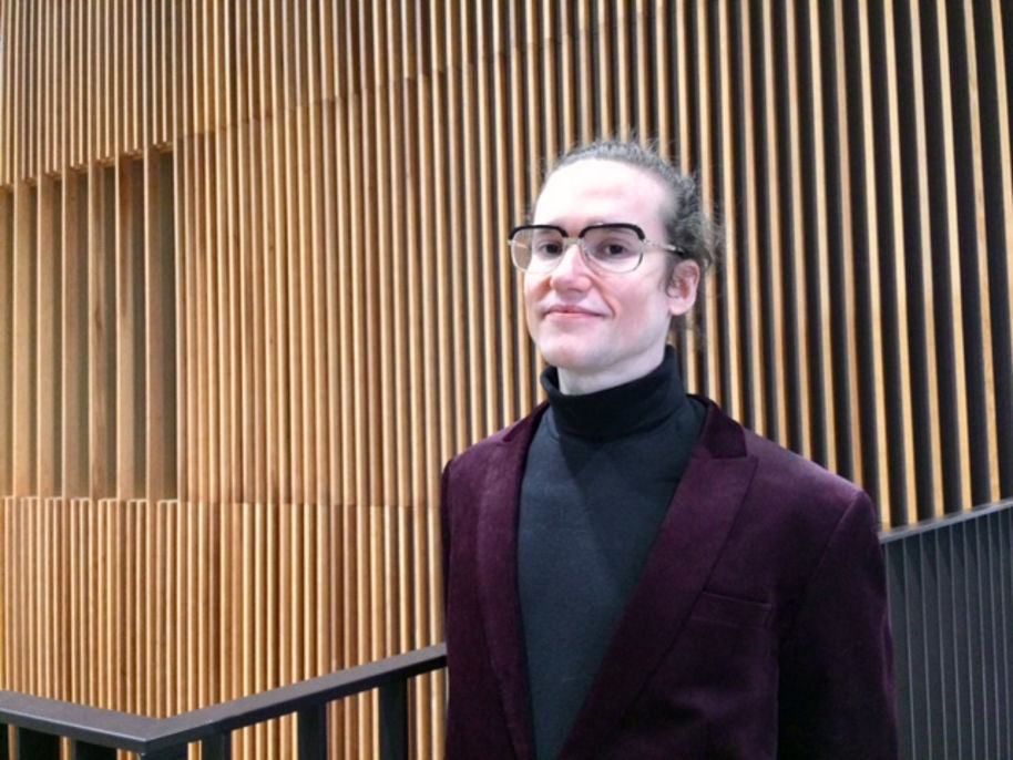 silmälasipäinen mies seisoo puurimoista tehdyn seinän edessä yllään violetti pikkutakki