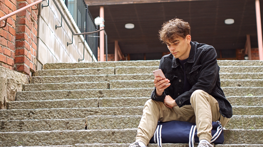 Nuori mies istuu rakennuksen kiviportailla ja selailee puhelintaan.