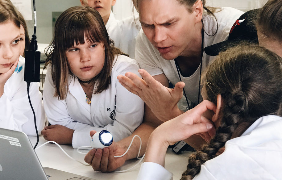 Lapsiryhmä tutkii ohjaajan kättä USB-mikroskoopilla. Lapsilla on laboratoriotakit.