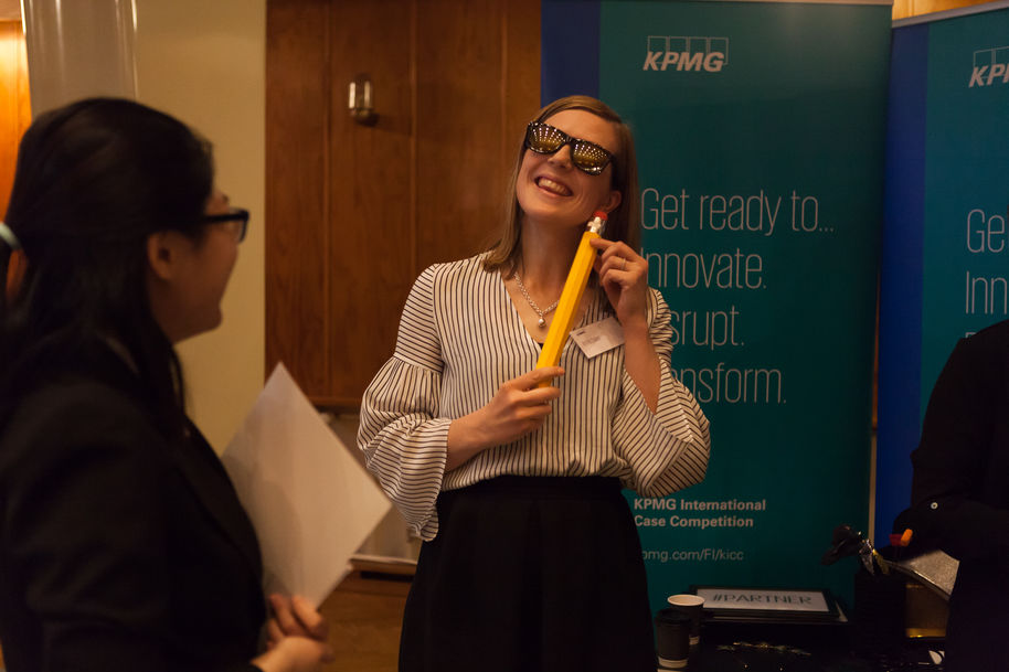 A woman is standing in front of roll-ups with KPMG logo, wearing sunglasses, smiling and holding a large pen next to her face.