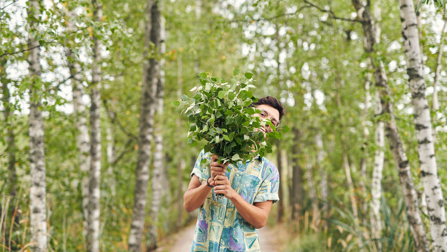 A person cheerfully peeks out from behind the fresh wind in a summery, green birch landscape.