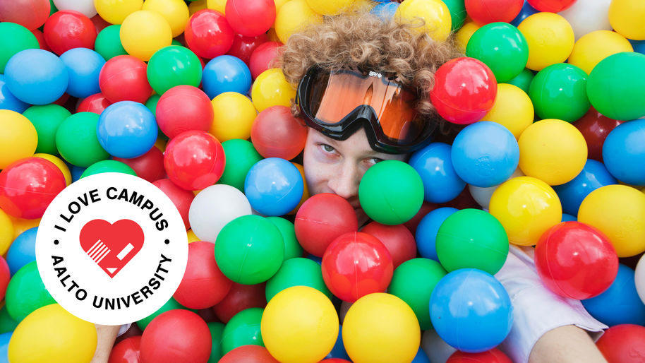 A person peeks through colourful balls in a ball pit. I love campus logo badge in the lower left corner.