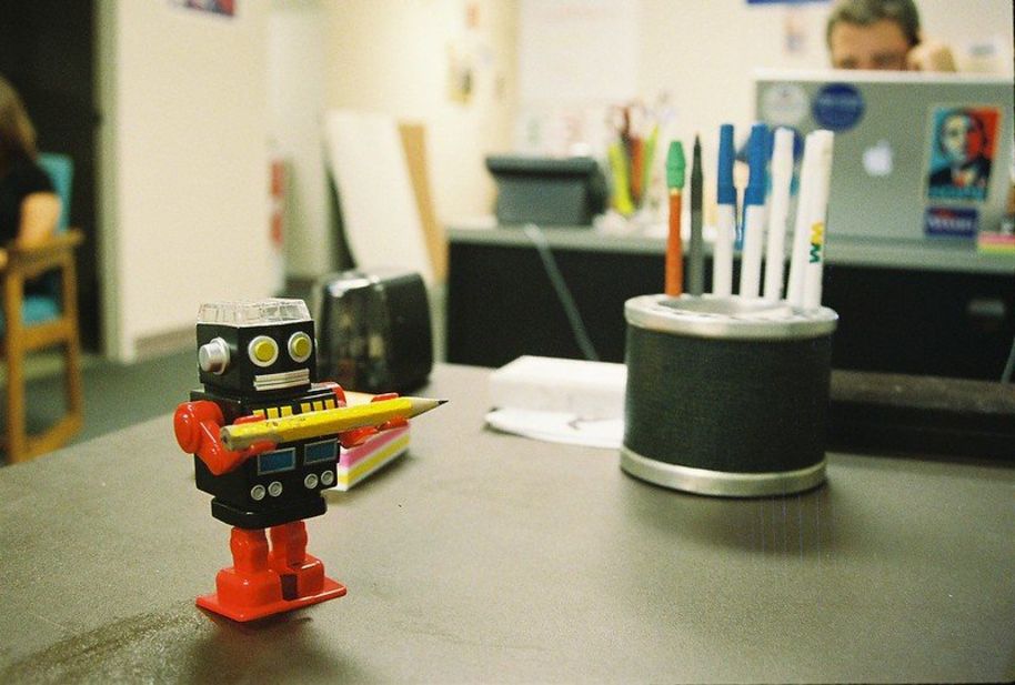 Red and black cartoonish robot with yellow eyes on a table, carrying a pencil against a classroom backdrop.