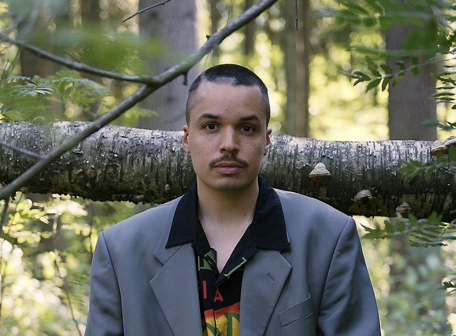 lyhyttukkainen mies seisoo metsässä ja katsoo suoraan kameraan, harmaa puvuntakki ja monivärinen paita
