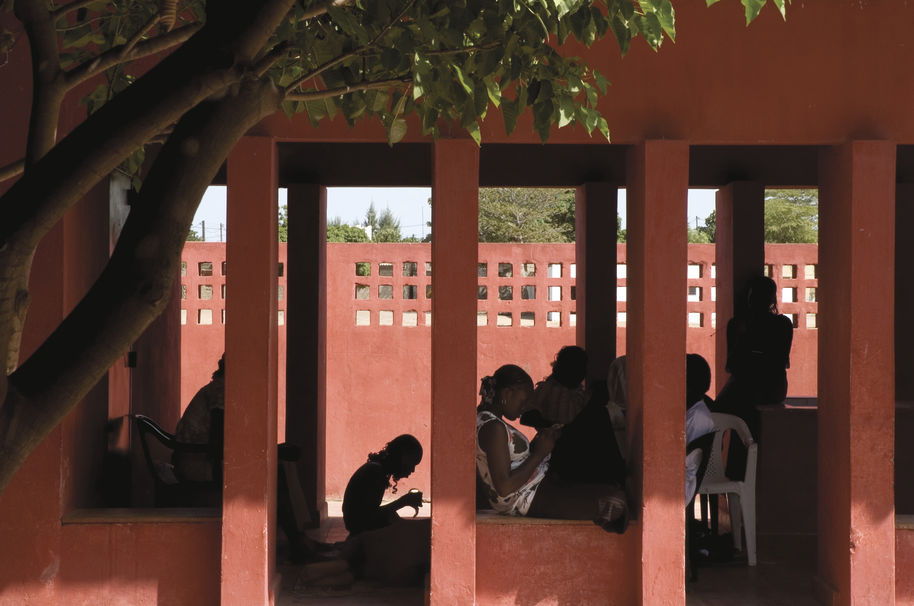 Nuoria afrikkalaisia naisia istumassa ja lukemassa punasävyisen katetun terassirakennuksen suojissa. Etualalla on puu.
