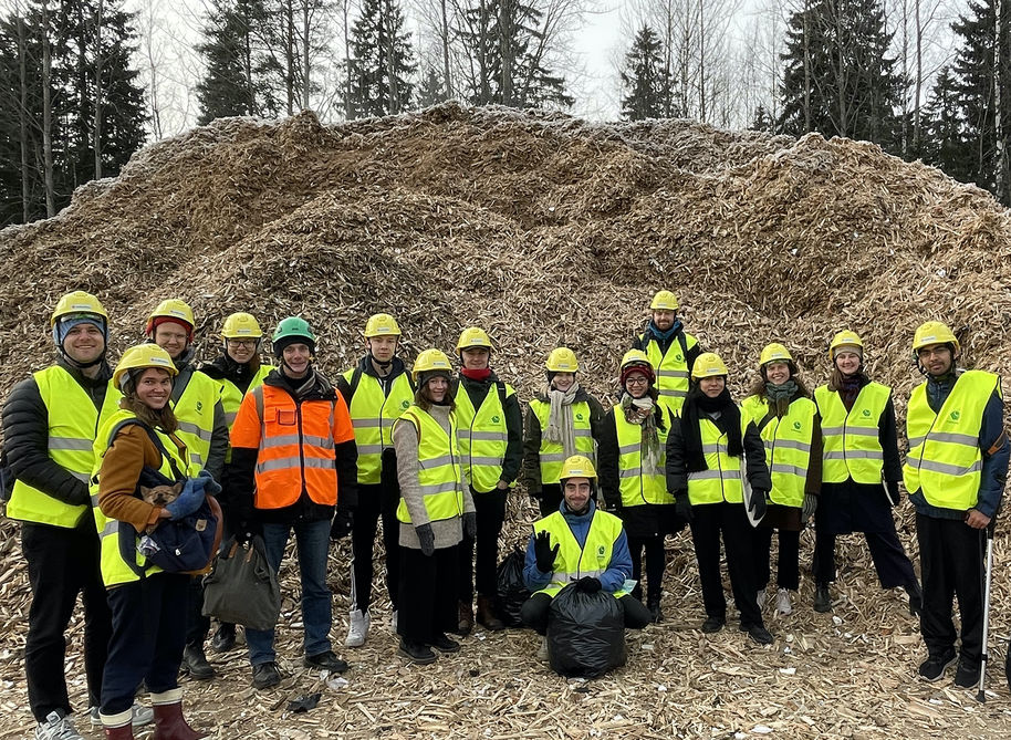 Sixteen students and teachers standing in front of a pile of wood chips in yellow and orange safety gear