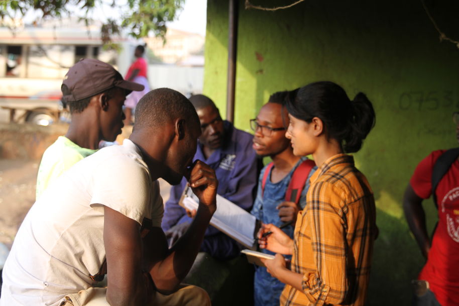 Photograph shows two graduate students with clipboards and notebooks talking with three local people in Uganda