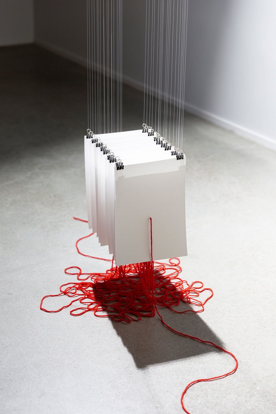 Installation made up of hanging pieces of white paper being pierced by a long, red string that piles up underneath.