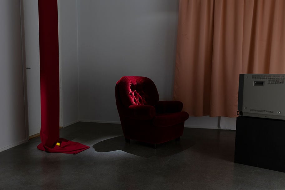Installation view. A red velvet armchair is illuminated from above. A red piece of fabric hanging from the ceiling and piling up on the ground, where a small yellow ball sits on top.
