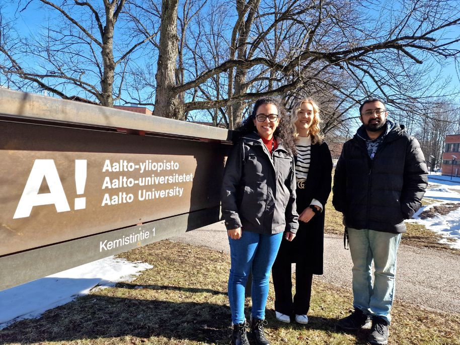 Colour photo of three smiling people standing outside under trees next to a sign that reads Aalto University, Kemistintie 1. There is snow on the ground and it is sunny.