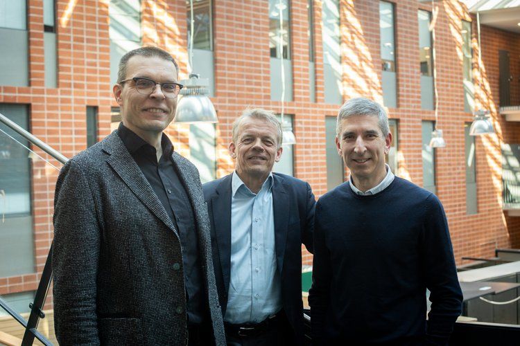 Three male researchers in dark smart casual clothing posing for a photo in front of a red-brick wall