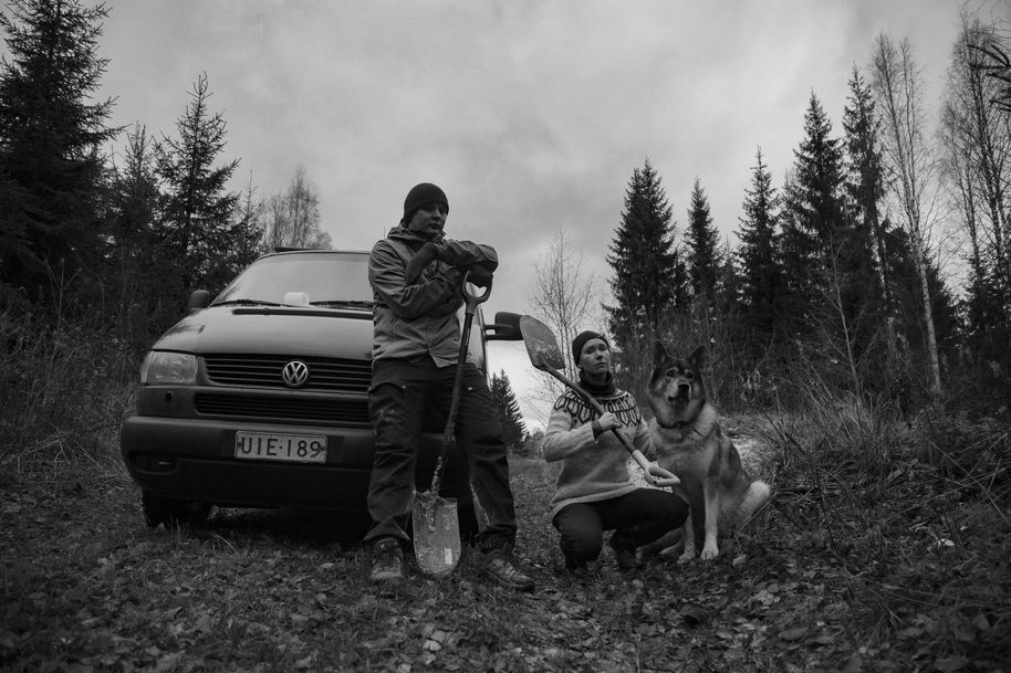 A black a white photo of Mira, her dog and a man standing in front of a car in a forest, holding shovels, posing for a picture before they dig up some wild clay