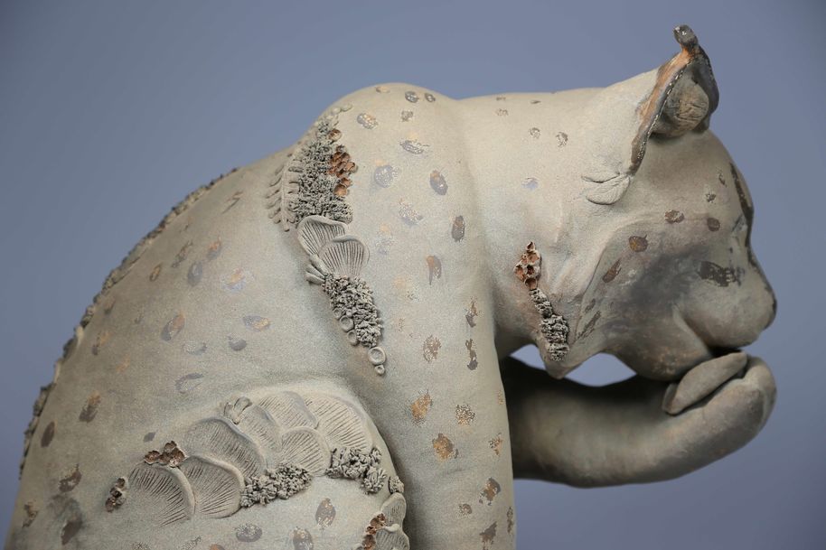 A ceramic sculpture of a lynx holding its paw close to its snout, there are mushrooms and lichen growing out of the lynx