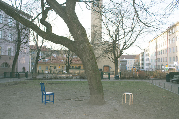 A large tree in a residential square leaning to the left, on the left of iit is a blue chair and on the right a wooden stool