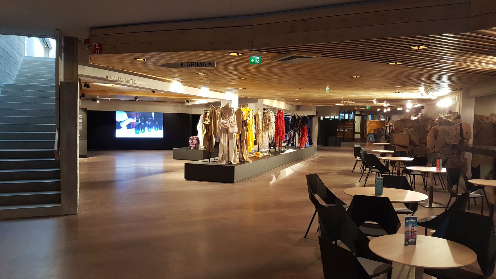 Dipoli-rakennuksen pääsisäänkäynti nimeltään Käpyaula. Kuvassa pitkä aulakäytävä, missä kahvipöytiä ja muodin opiskelijoiden suunnittelemia ja valmistamia vaatteita. Kuva: Aalto-yliopisto, Mirka Elovaara 