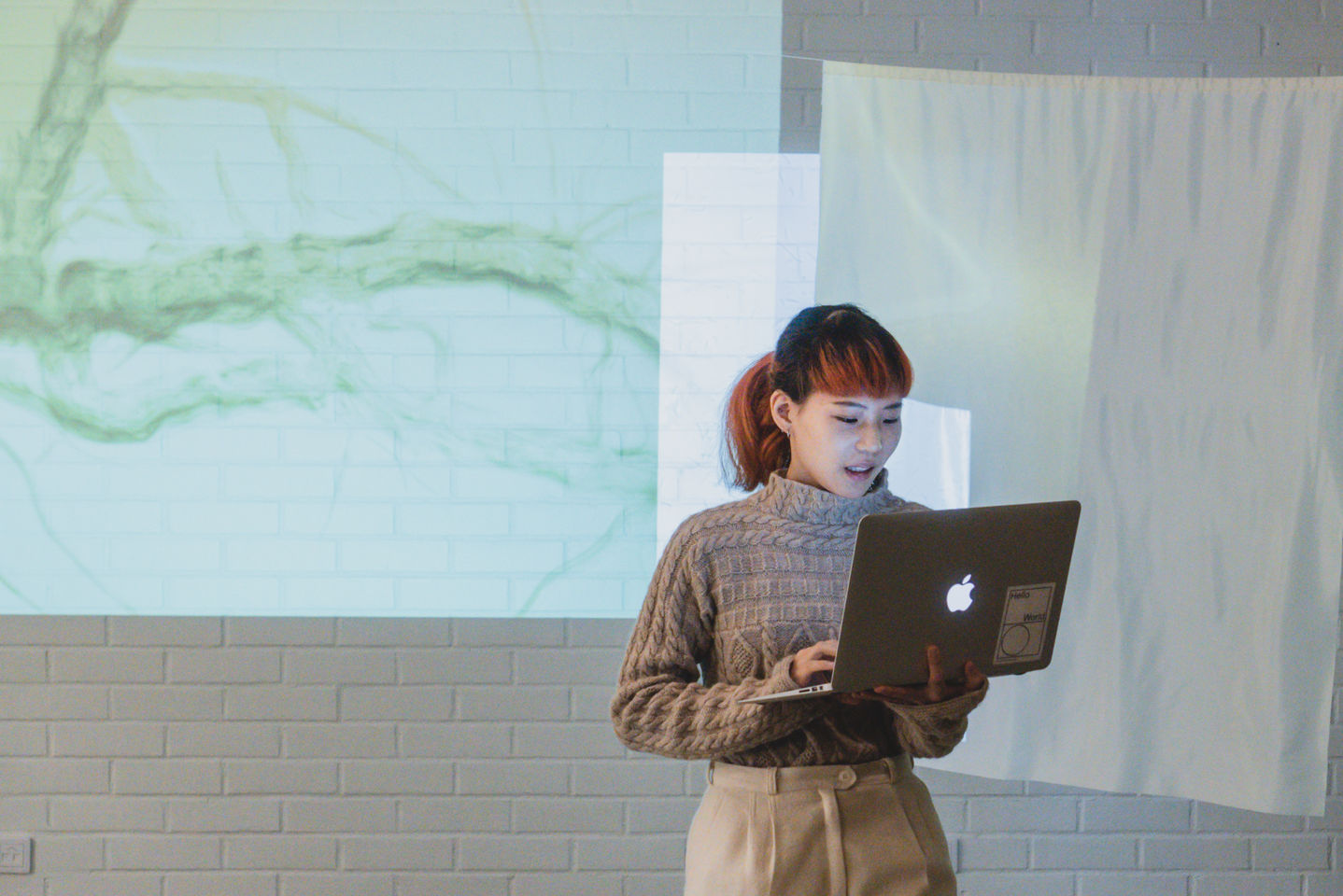 A ViCCA student stands in front of projected screens talking about her work with a macbook in hand