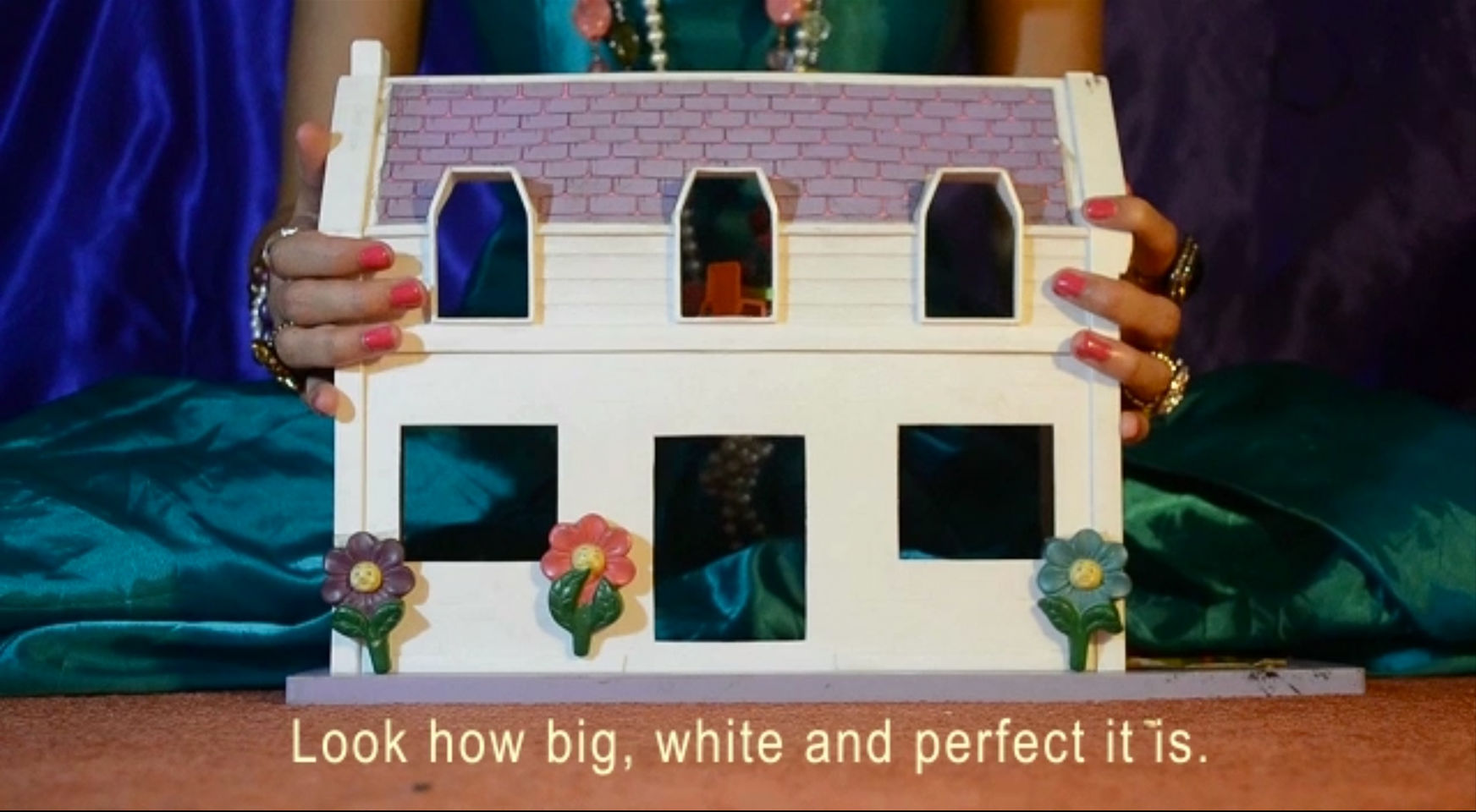 A video still featuring a white doll house with a woman sat behind it