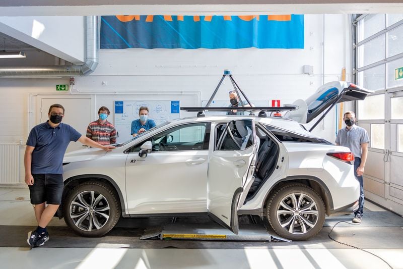 Autonominen A!ex-auto kuvattuna valoisassa autohallissa ympärillään viisi tutkijaa kesäisissä vaatteissa ja kasvoillaan maskit. Valkoisen auton katolla on tekninen laite ja auton tavaratilan kansi on ylhäällä.