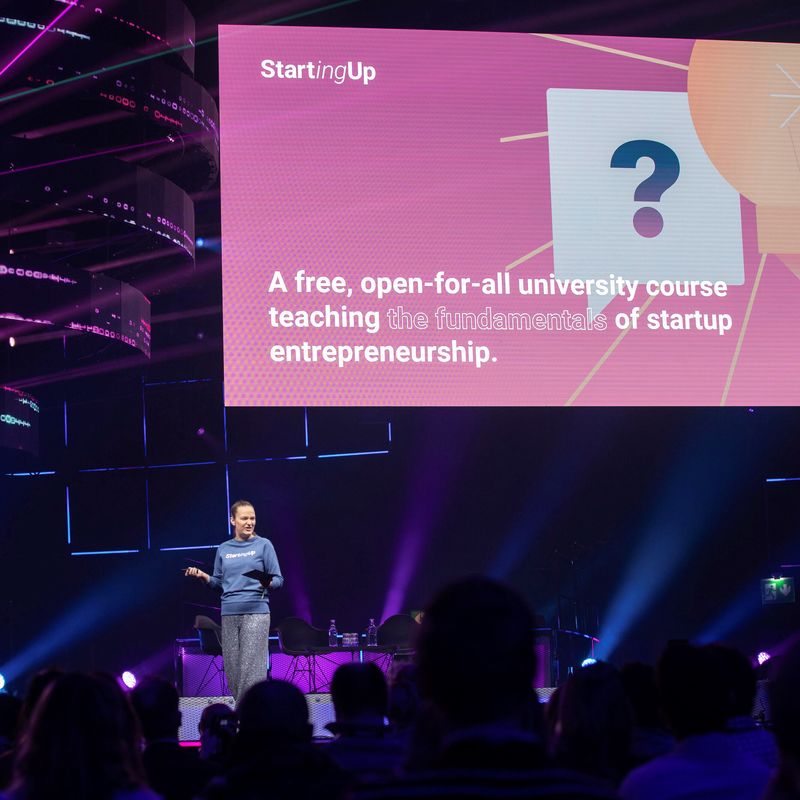 A woman stands on stage at Slush, presenting Starting Up online course, lights are purple