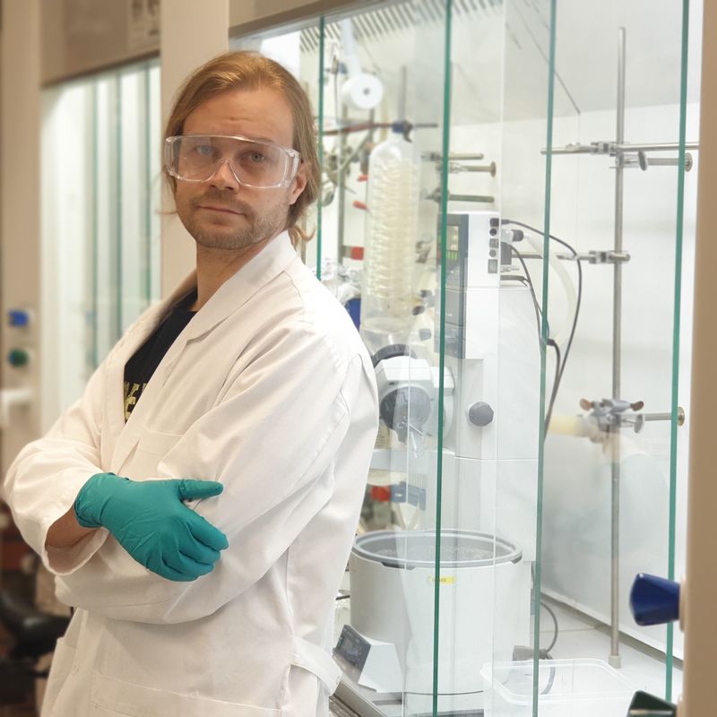 Apulaisprofessori Juha Siitonen laboratoriossa, yllään laboratoriotakki, suojalasit ja -hanskat.